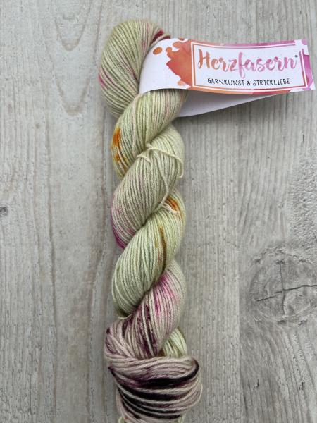 Ein handgefärbter Strang Herzfasern No. 94 – Cashmere Cloud in der Farbe Blütenstaub. Sanfte Creme- und Rosétöne mit Akzenten in Goldgelb, Pink und Bordeaux. Der Strang liegt auf hellem Holz, mit Herzfasern-Banderole.